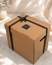 Load image into Gallery viewer, Brown cardboard box with a black ribbon and label on a light surface.
